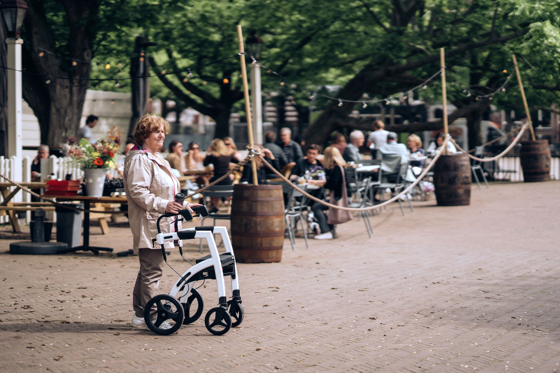 A woman with a rollator pausing beside a tree-lined café terrace